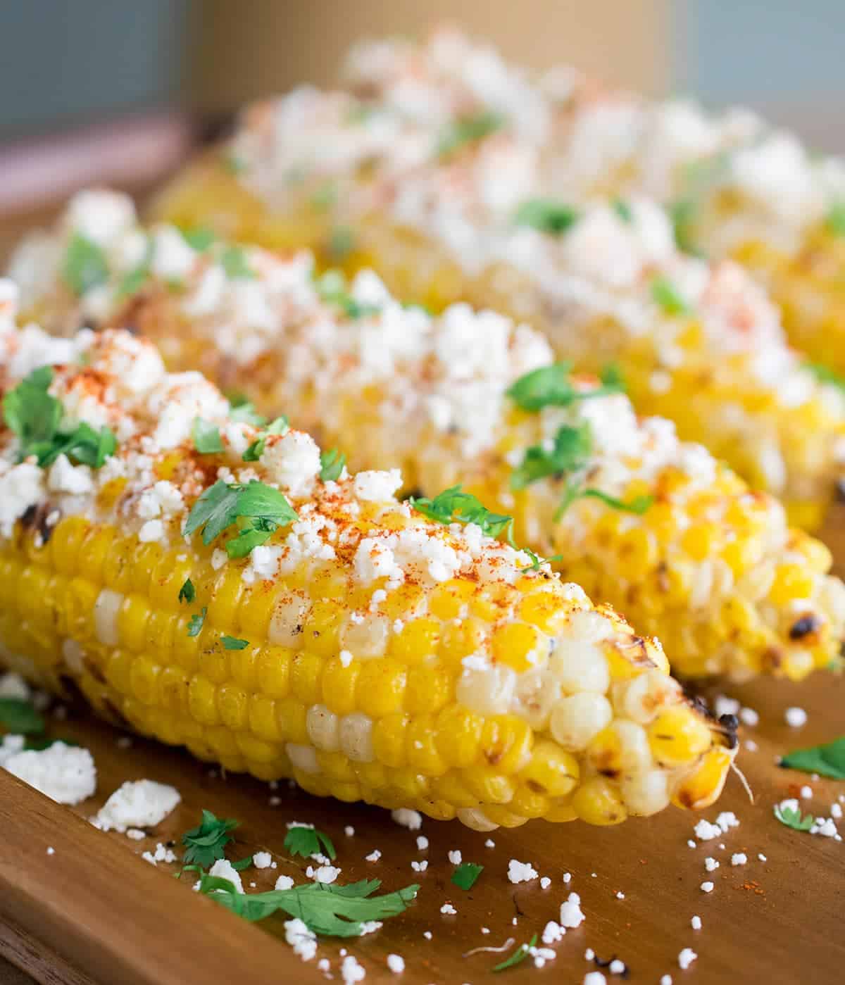 Mexican street corn topped with cotija cheese, cilantro & chili powder on wooden cutting board.