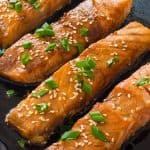 Close up of four pieces baked Salmon in a pan in Teriyaki sauce with sesame seeds and chives garnish.