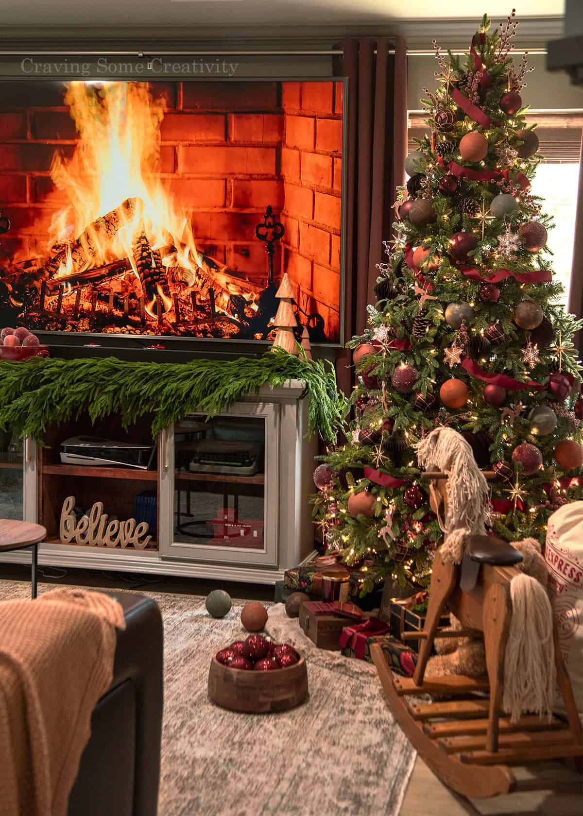 A Christmas living room scene with Victorian Christmas tree, fireplace TV, and red decorations all around.