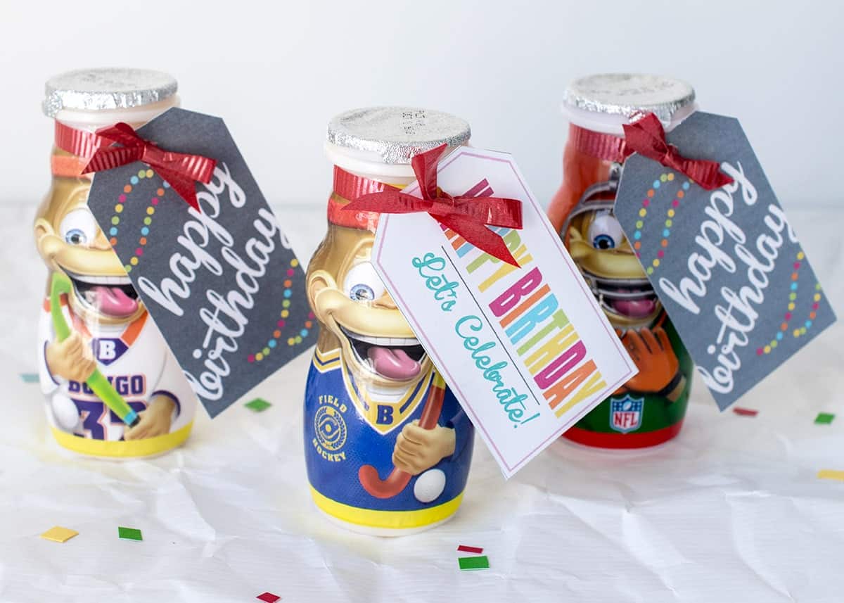 Classroom birthday treats-yogurt smoothie treats wrapped in red bows with printable labels on white surface with colored confetti.