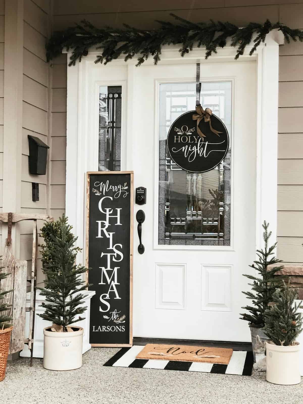 Black and white holiday decorated front door with small trees in pots and a DIY sign and hanging door sign.