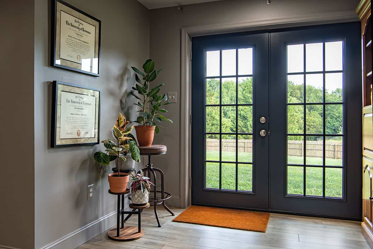 Black French doors in a medium gray room with plants on stands, cabinetry and artwork.