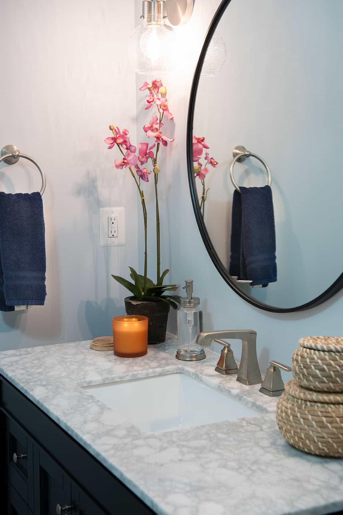 Marble bathroom vanity with round mirror and decor such as a candle and orchid.