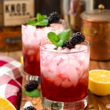 Two glasses of blackberry bourbon smash cocktail garnished with mint and blackberries on a wood table.