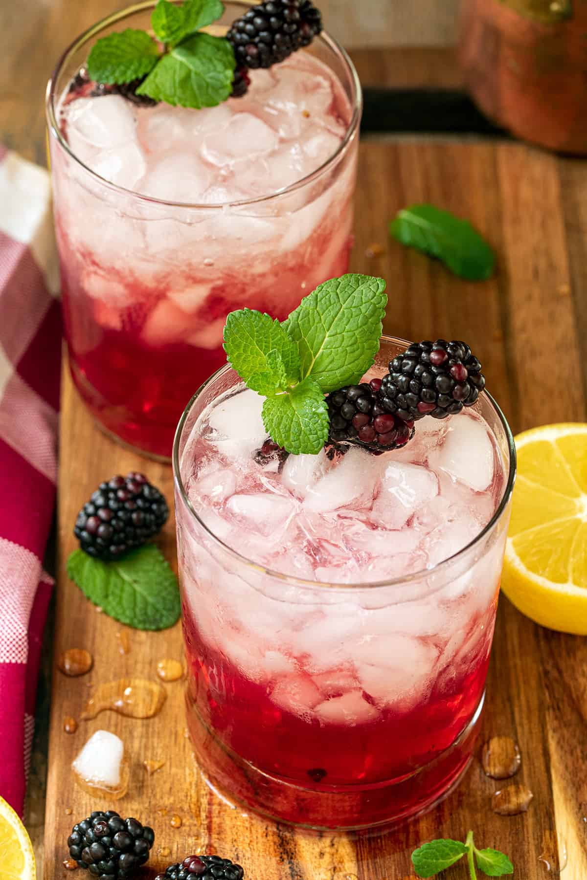 Two glasses top down view of blackberry bourbon cocktails with sprig of mint and blackberries as garnish on a wooden tray.