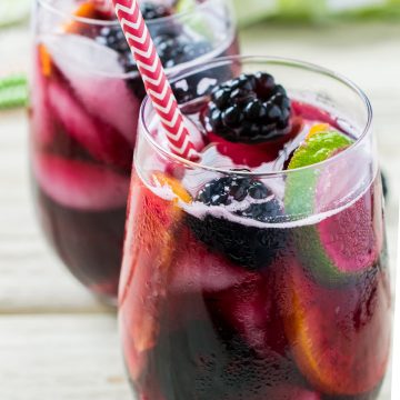 Two glasses of blackberry sangria with straws on a wood background.