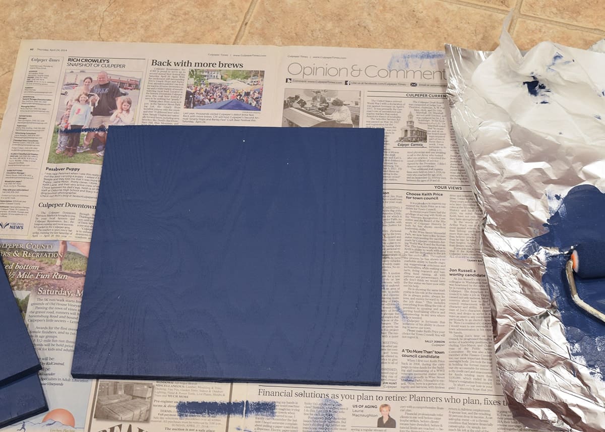 Newspaper with freshly painted navy blue wood square on top next to aluminum foil covered with paint.