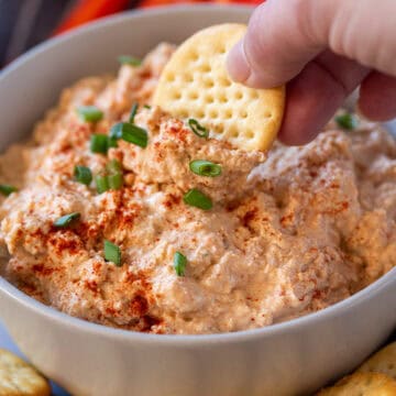 A butter cracker being dipped into cold Cajun crab dip with green onions on top.