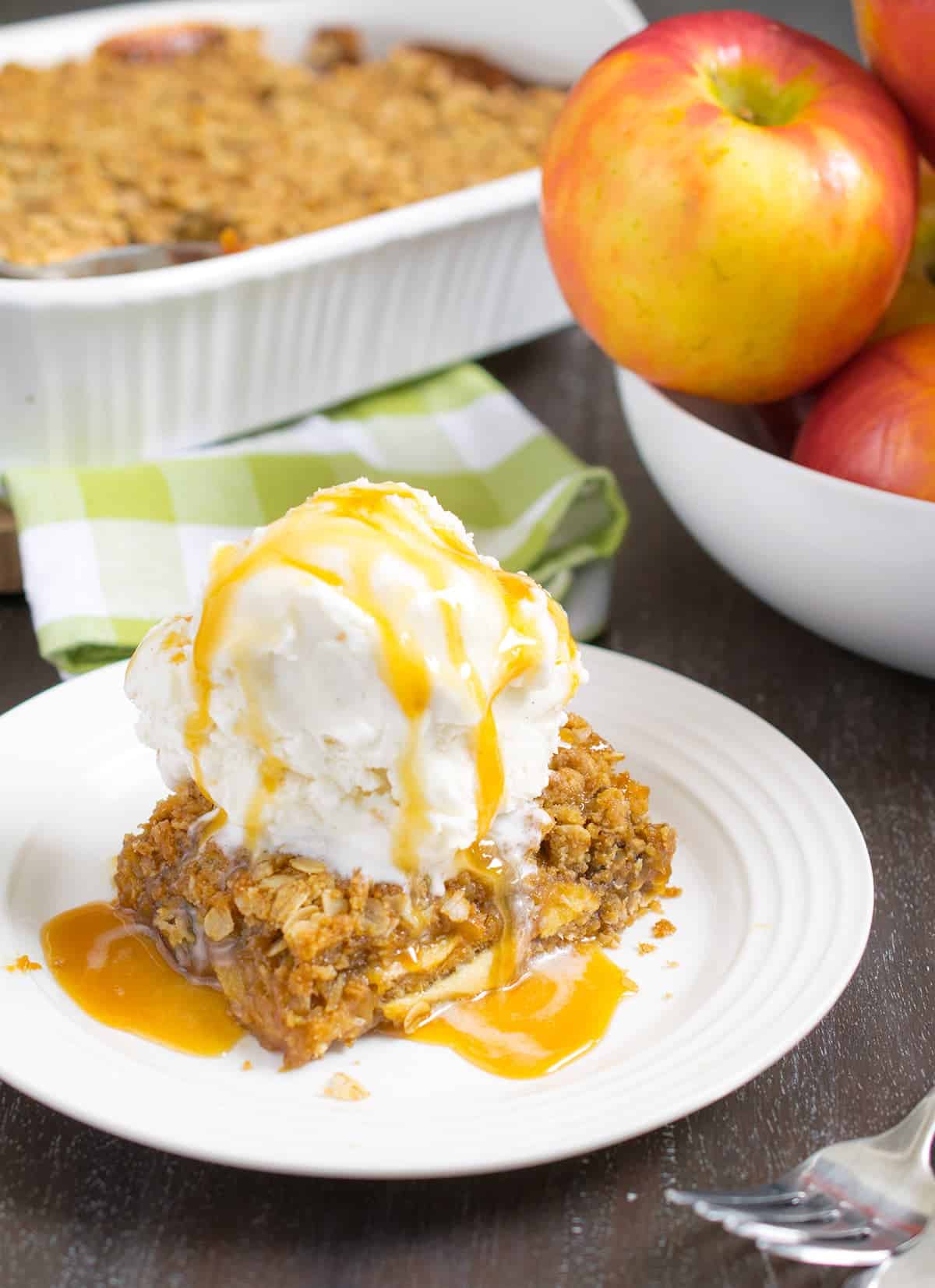 Baked caramel apple crumb bars with vanilla ice cream and caramel sauce on dessert plate next to a bowl of apples.