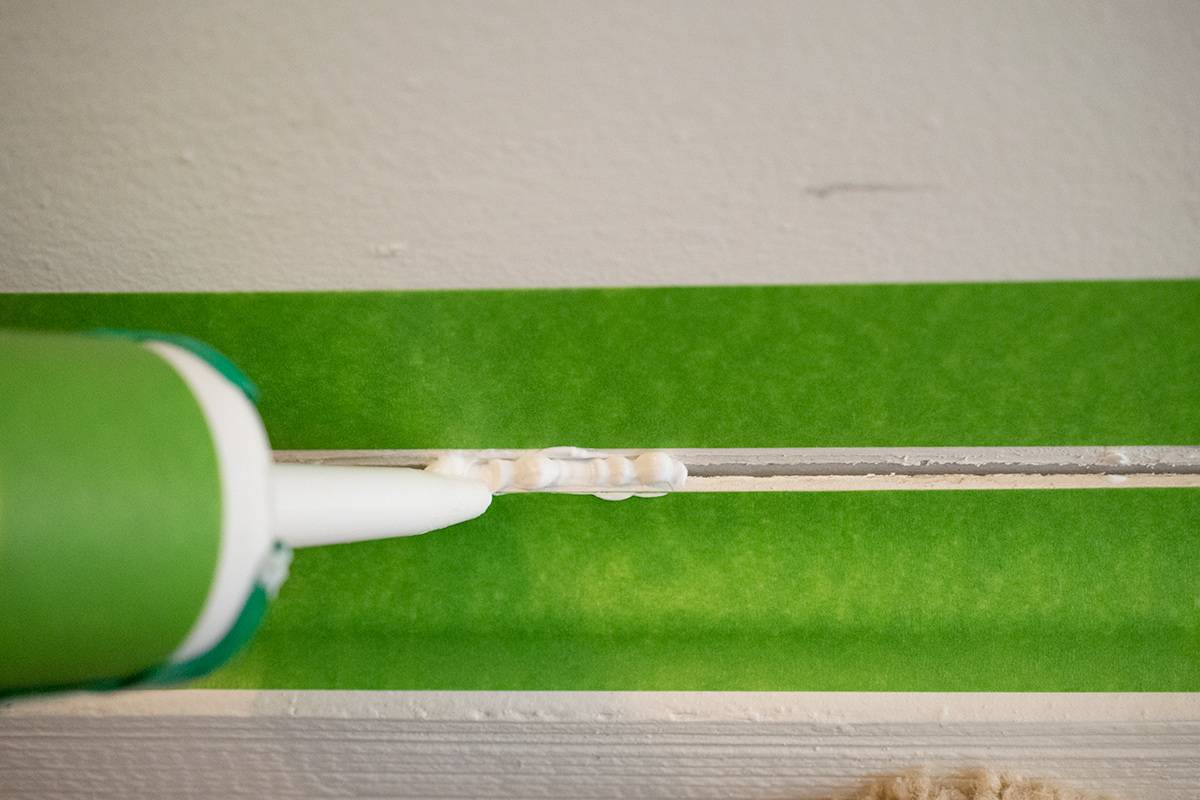 Caulking gun applying caulk to baseboard between strips of green frog tape.