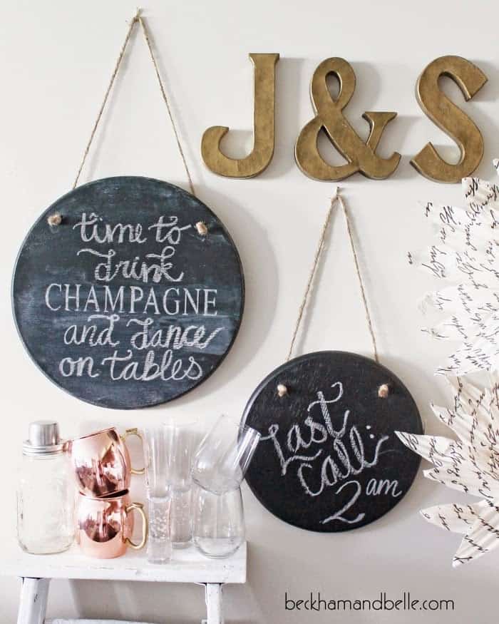 Round hanging wood wall chalkboard art above white side table with assorted glasses and bronze letter art.
