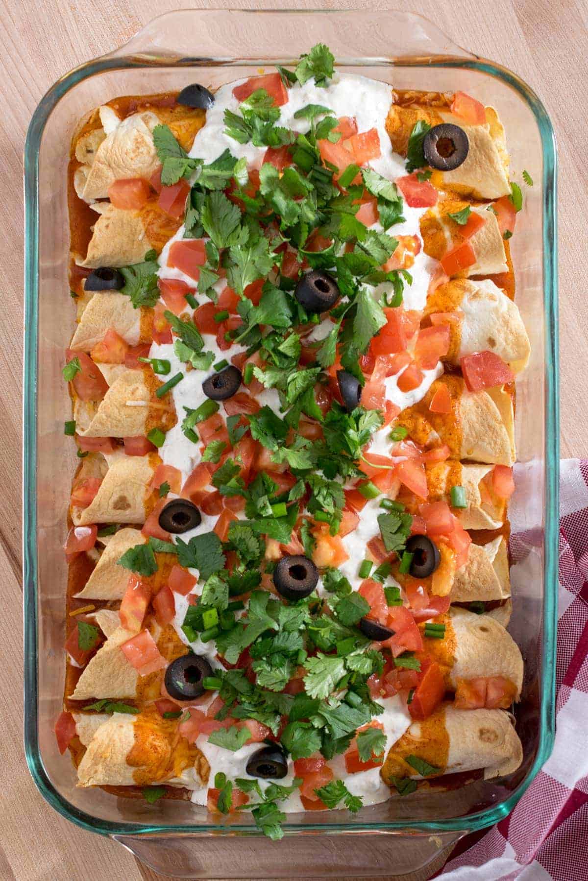 Top down view of baked chicken enchiladas with sour cream, pico de gallo, olives, and cilantro as garnish in glass baking dish.