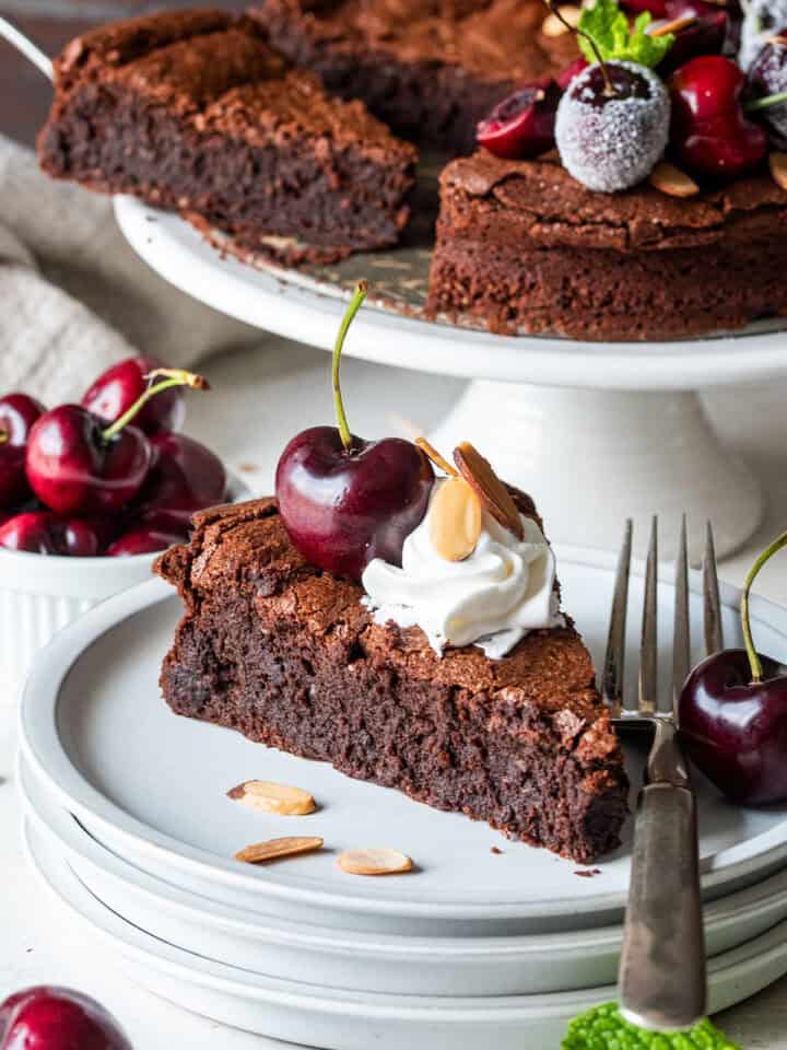 A slice of flourless chocolate almond cake with whipped cream on top in front of a full chocolate cake with cherries.