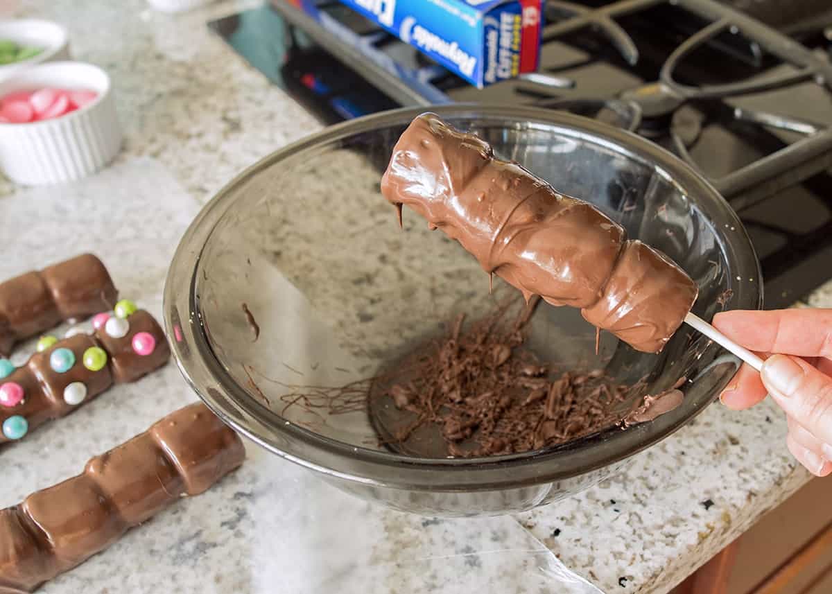 Excess dipping chocolate coating being shaken off of a marshmallow pop over a bowl.