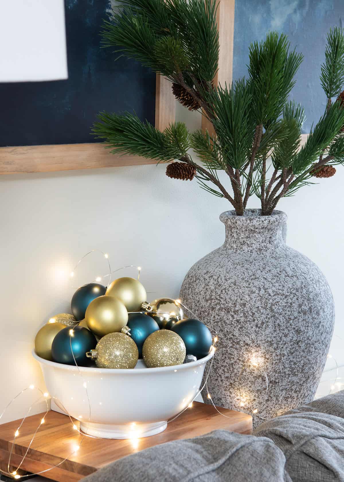 Vase of pine needles and a large bowl of ornament filler with twinkling fairy lights behind a sofa.