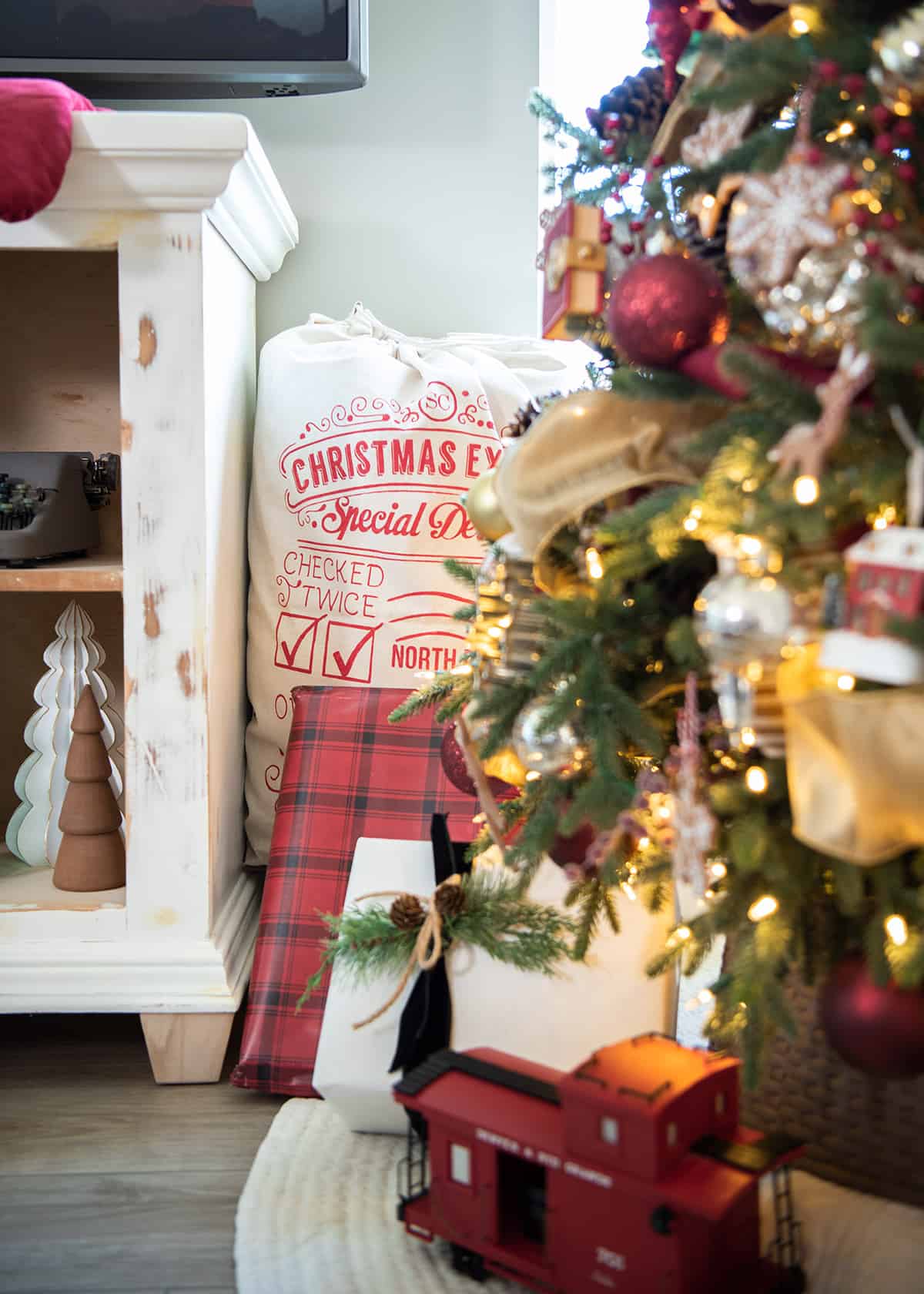 Santa sack tucked behind a red Christmas tree with plaid packages and a train underneath.