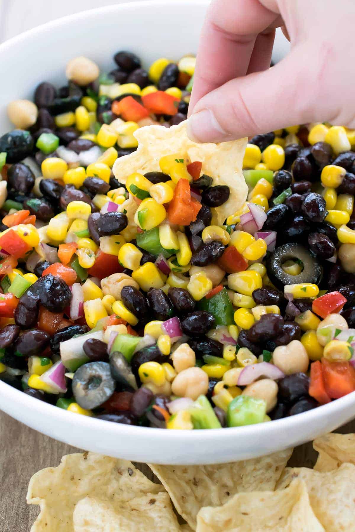 Light and fresh Cilantro black bean and corn salsa in bowl while a hand reahes in to scoop with tortilla chip.