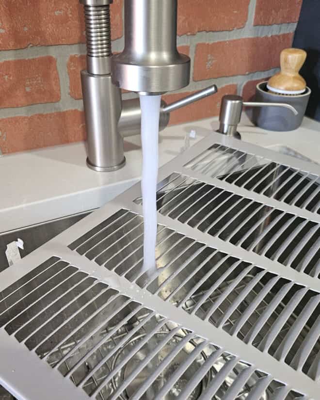 Washing an air return cover in the sink with hot water and a brush in the background.
