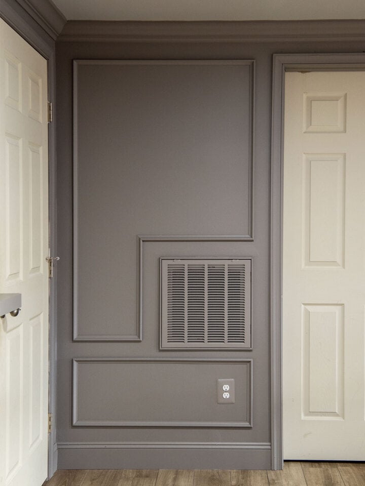 A grey wall with picture frame molding on it and a painted ventilation cover.