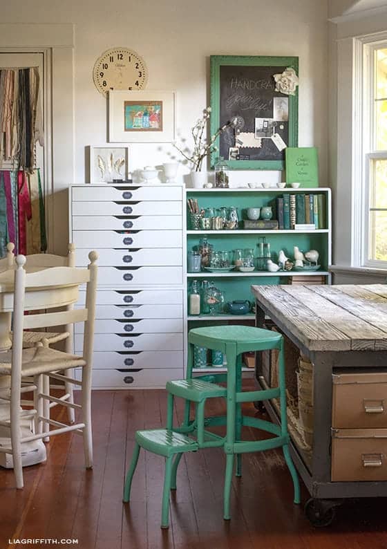 Vintage Craft room with rustic table, green accents, white cabinet, and collection of books and glass collectibles perfect for organizing your craft room efficiently.