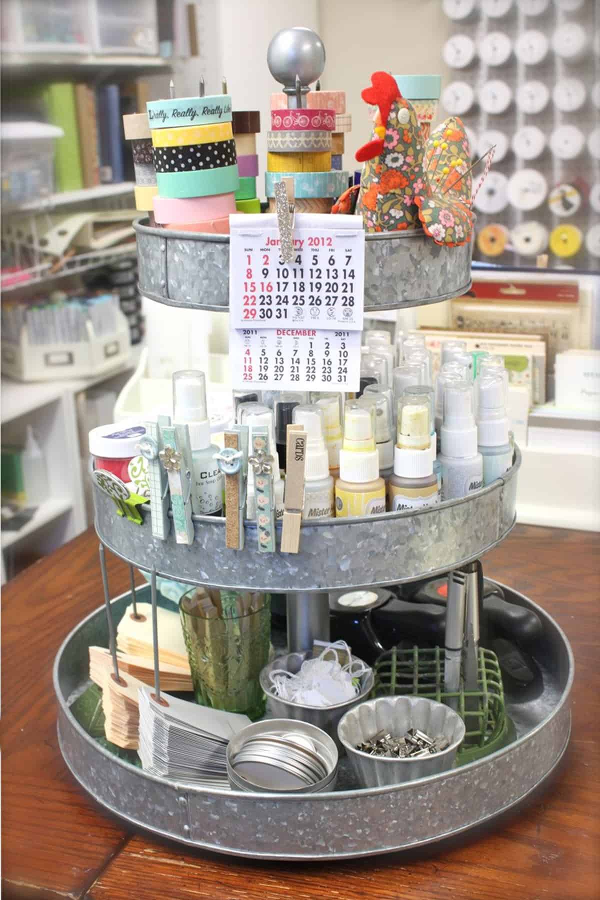 Metal tiered tray for organizing craft supplies with paints, ribbon spools, tags, clothes pins, and small calendar.