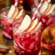 Apple cocktail with crown royal and pomegranate juice with apples and red striped straws for garnish.