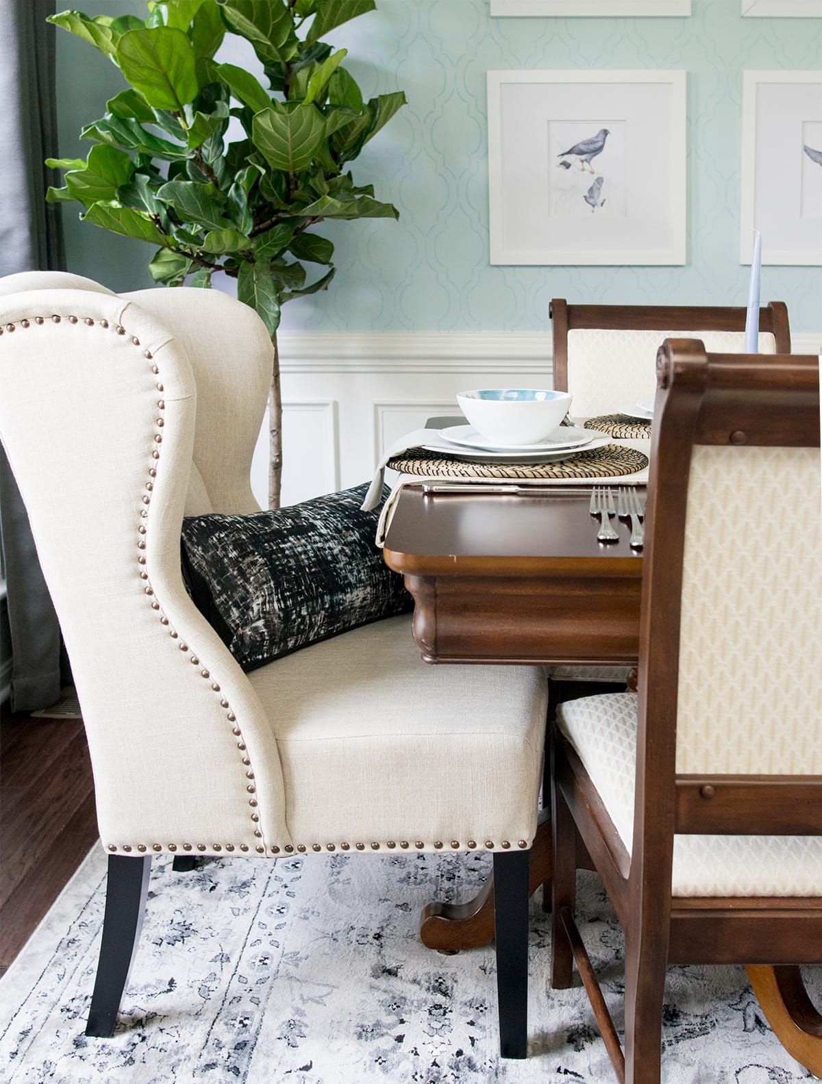 Studded white accent chair in dining room with pillow in front of potted plant and light blue wall with birdlife art.