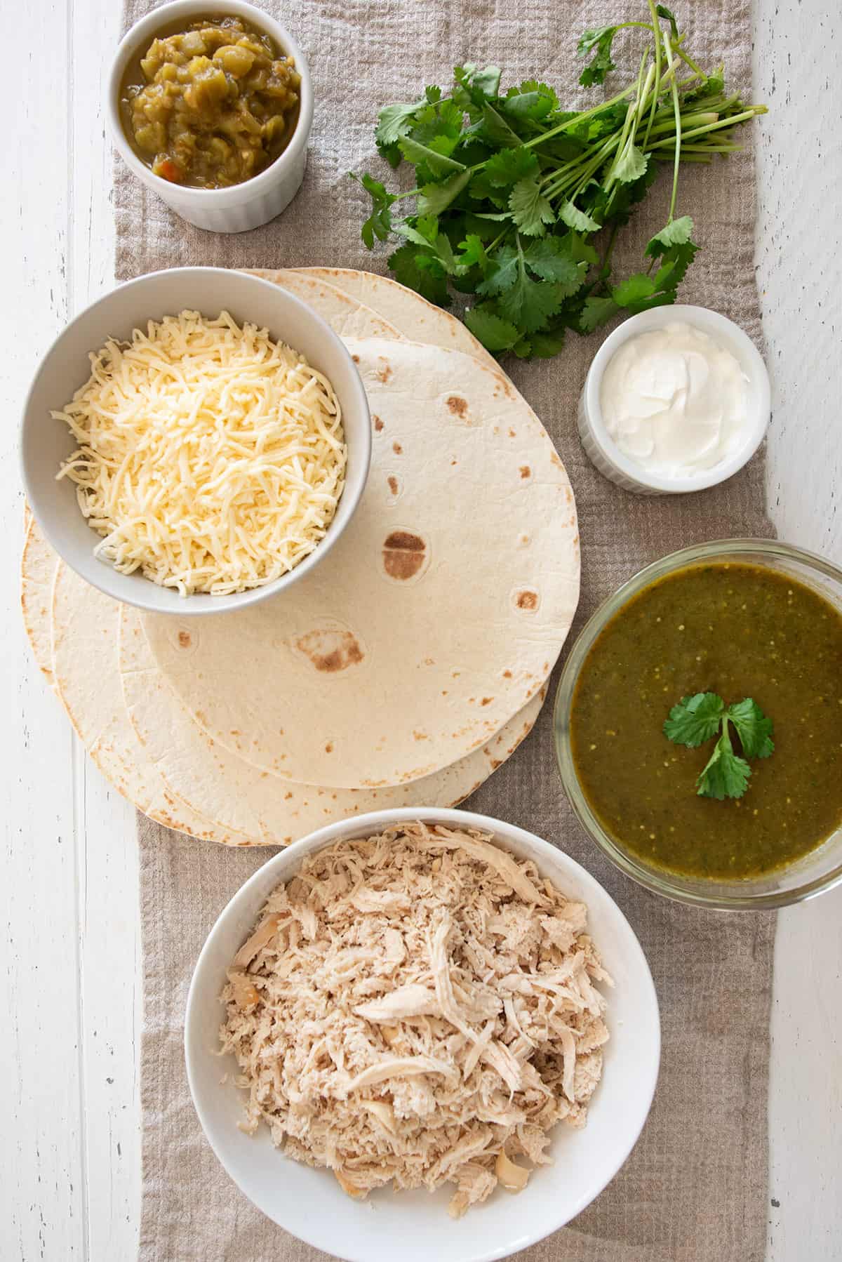 Enchilada filling ingredients laid out on a table such as tortillas, chicken, sour cream, green sauce, cheese, and cilantro.