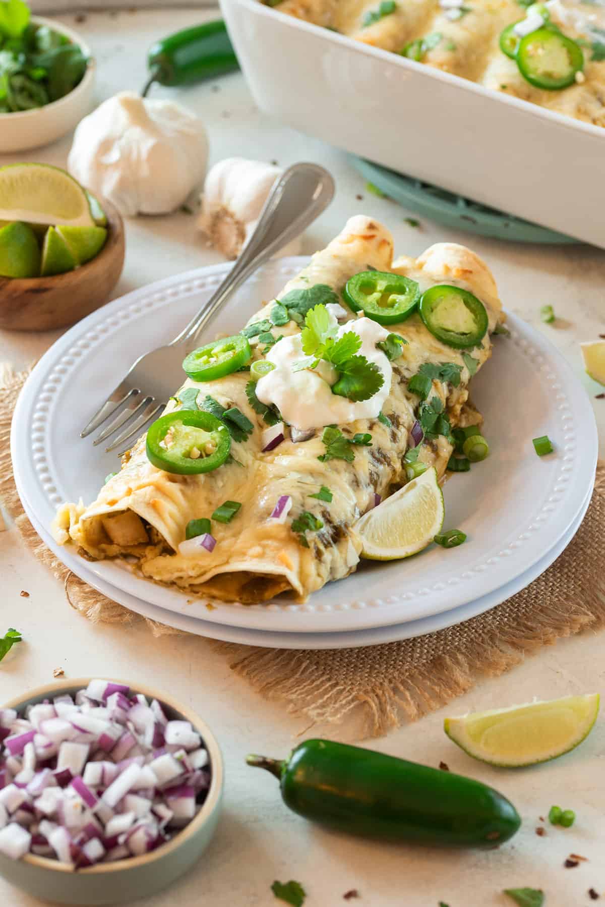 Two green enchiladas filled with chicken and topped with sour cream, cilantro, jalapeno and lime wedges on a plate.