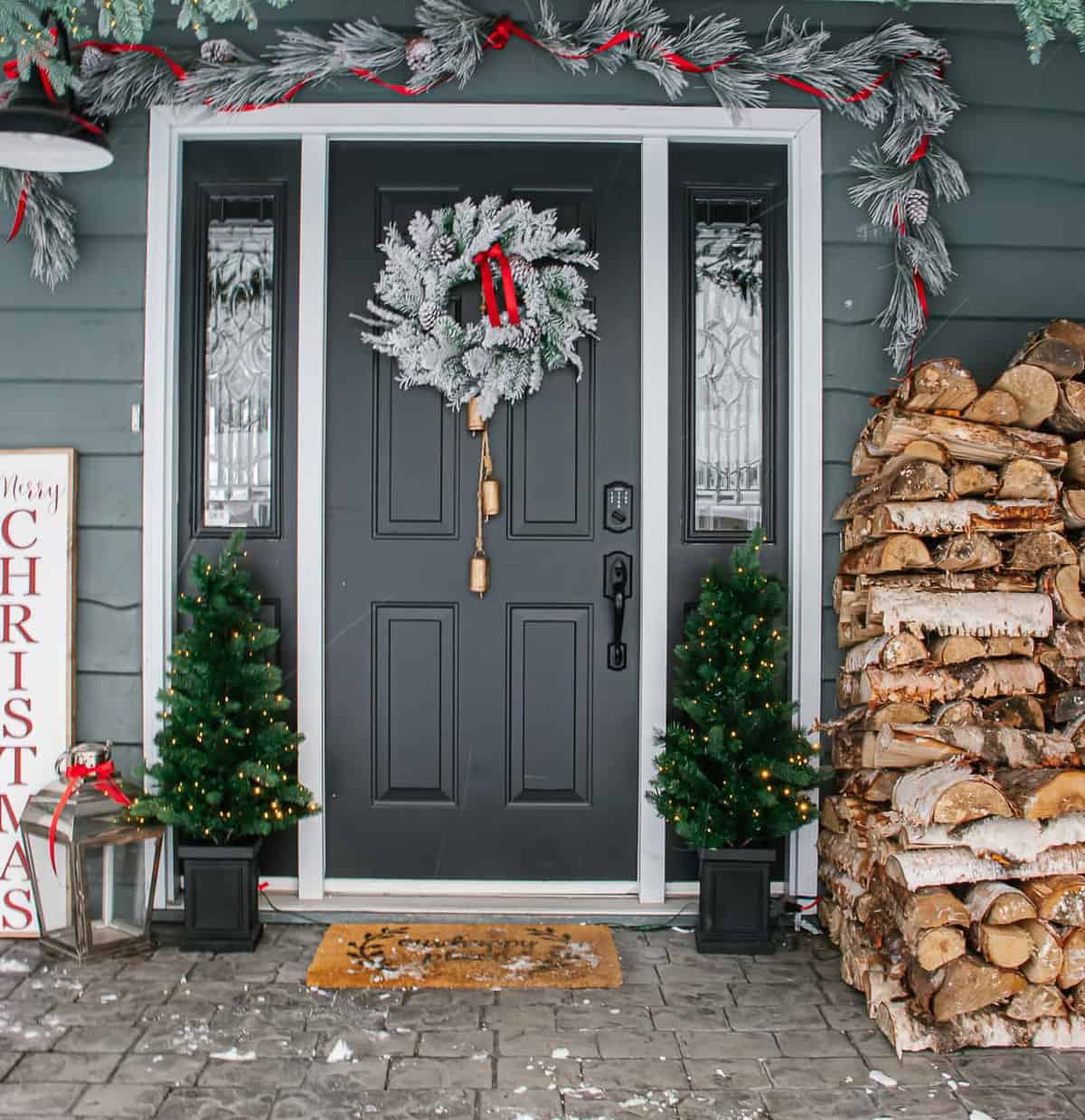 Wintry farmhouse Christmas porch with stacked firewood and flocked wreath and garland.