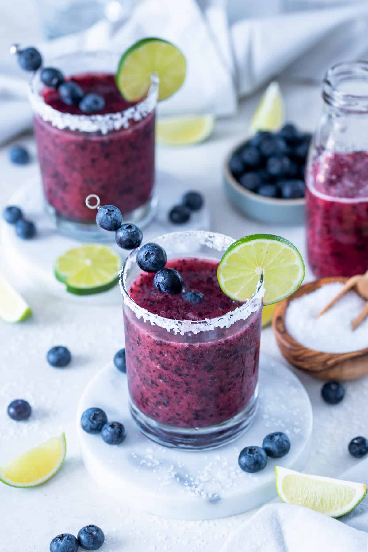 Two glasses of frozen blueberry margaritas with limes and blueberries sprinkled around a white background.