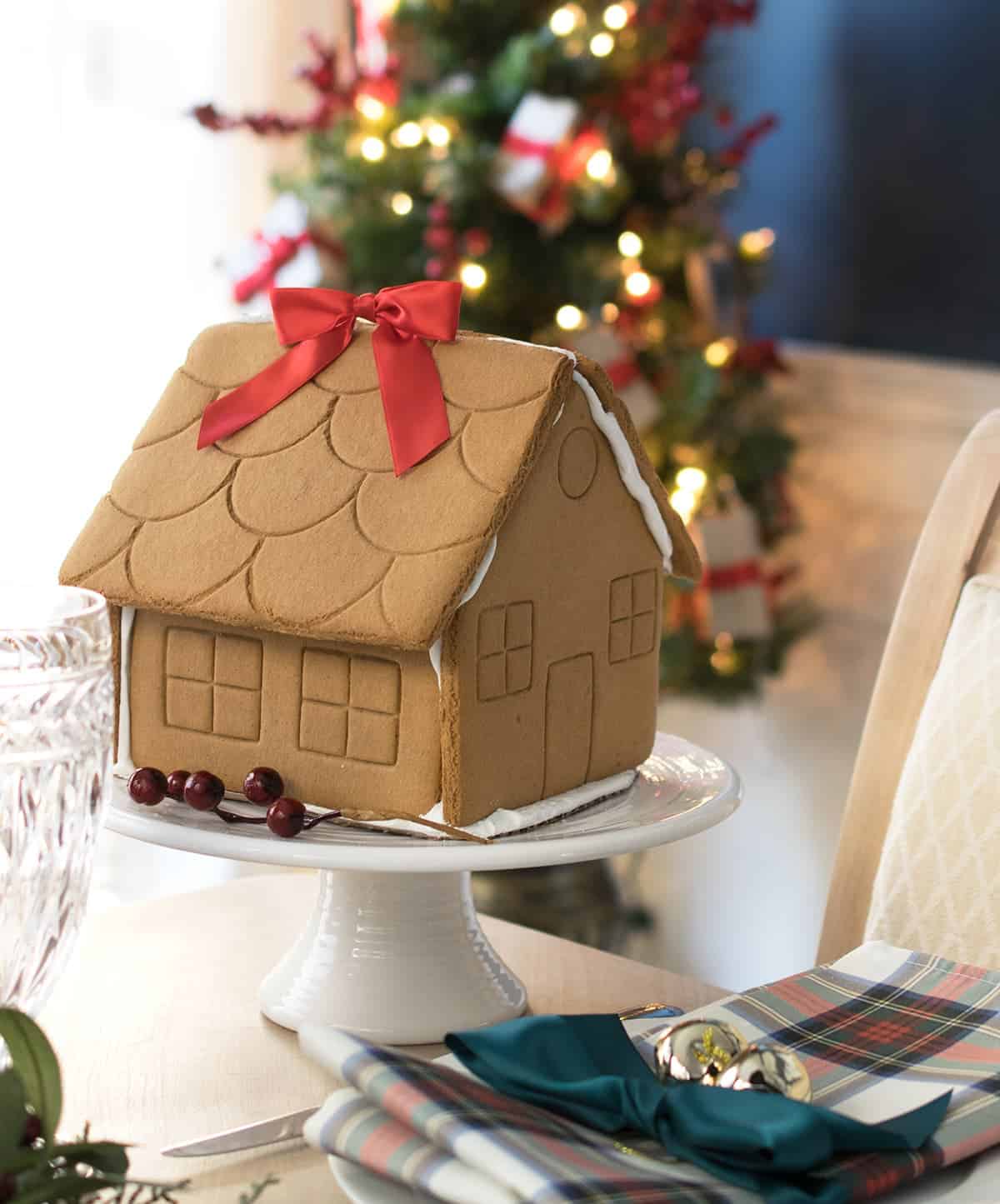 Christmas gingerbread house centerpiece on a white pedestal. Red ribbon and red berry sprig for accent.