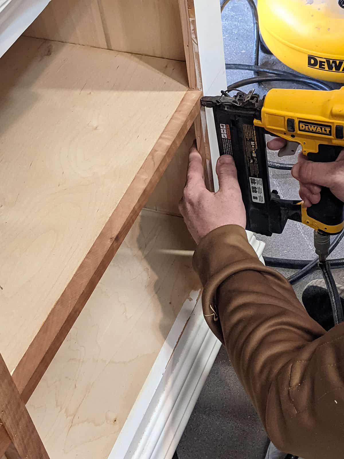 A man brad nailing pieces of wood to the front of plywood to conceal the rough edges like a veneer.