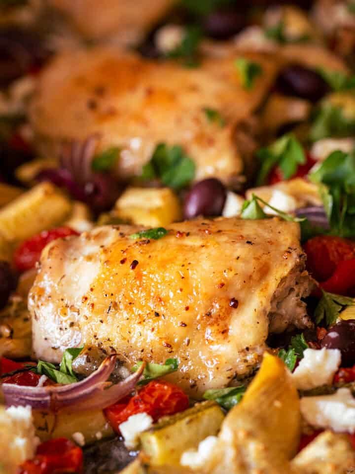 Closeup of a roasted chicken thigh nestled into a bed of cooked vegetables like tomato, red onion, olives, and artichokes.