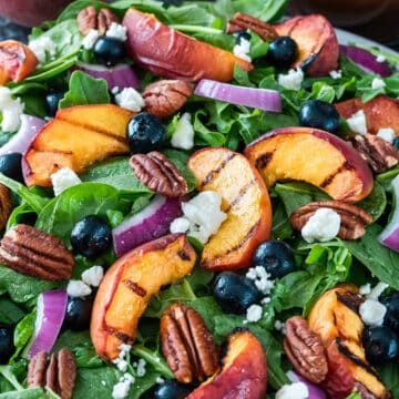 Spinach salad with grilled peaches, blueberries, cheese, pecans, and red onions on a plate.