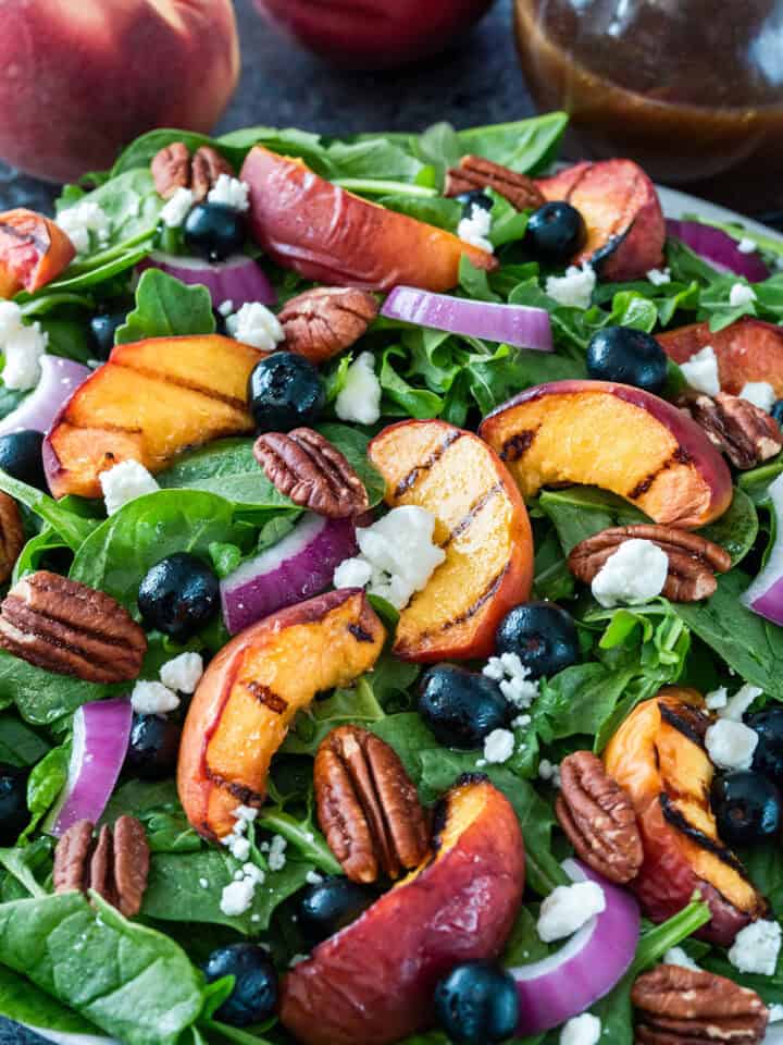 Spinach salad with grilled peaches, blueberries, cheese, pecans, and red onions on a plate.