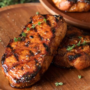 Two grilled pork chops with grill marks laid out on a wooden background.