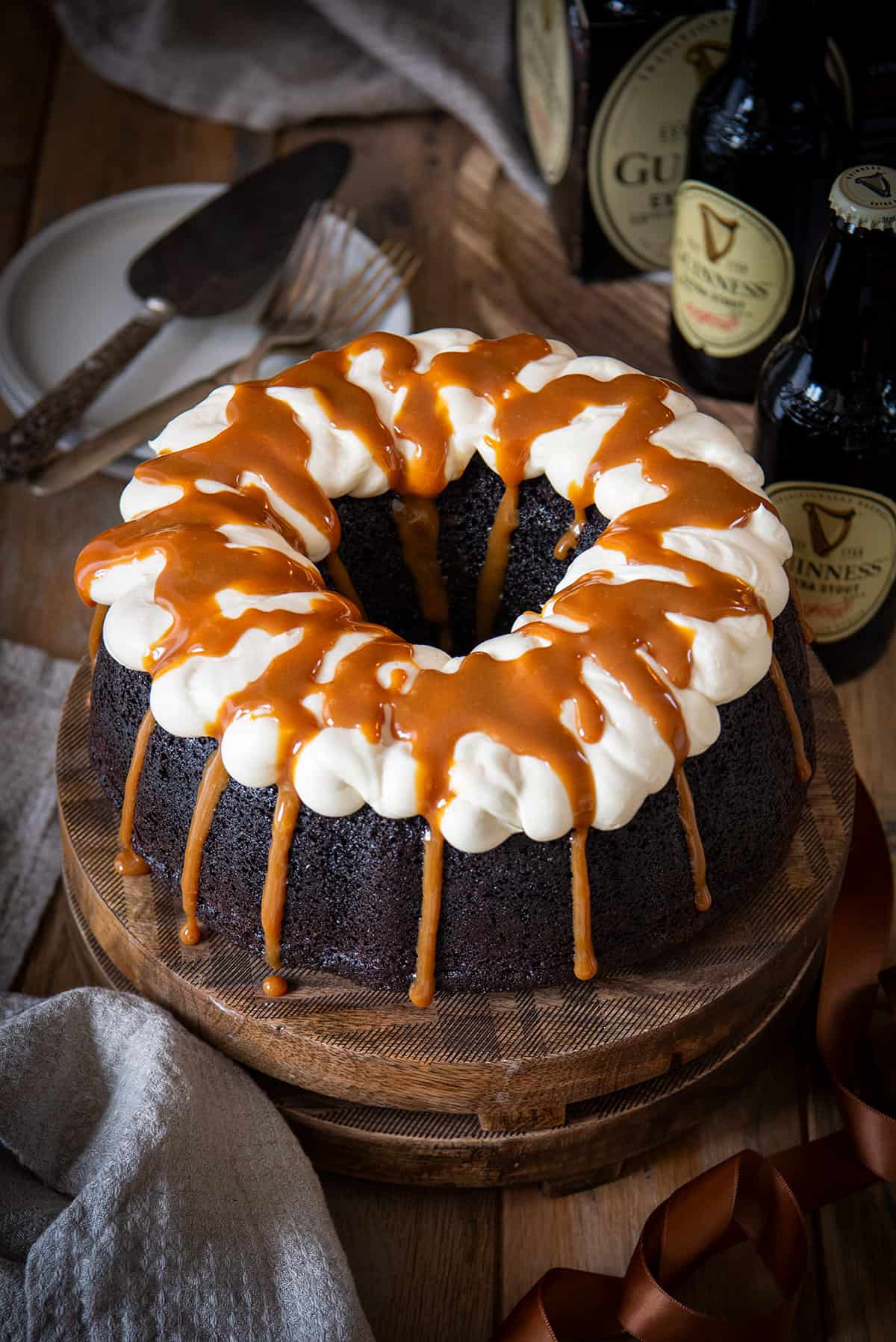 A Guinness Chocolate cake with vanilla buttercream frosting and a caramel drizzle on top in front of several Guinness bottles on a wood background.