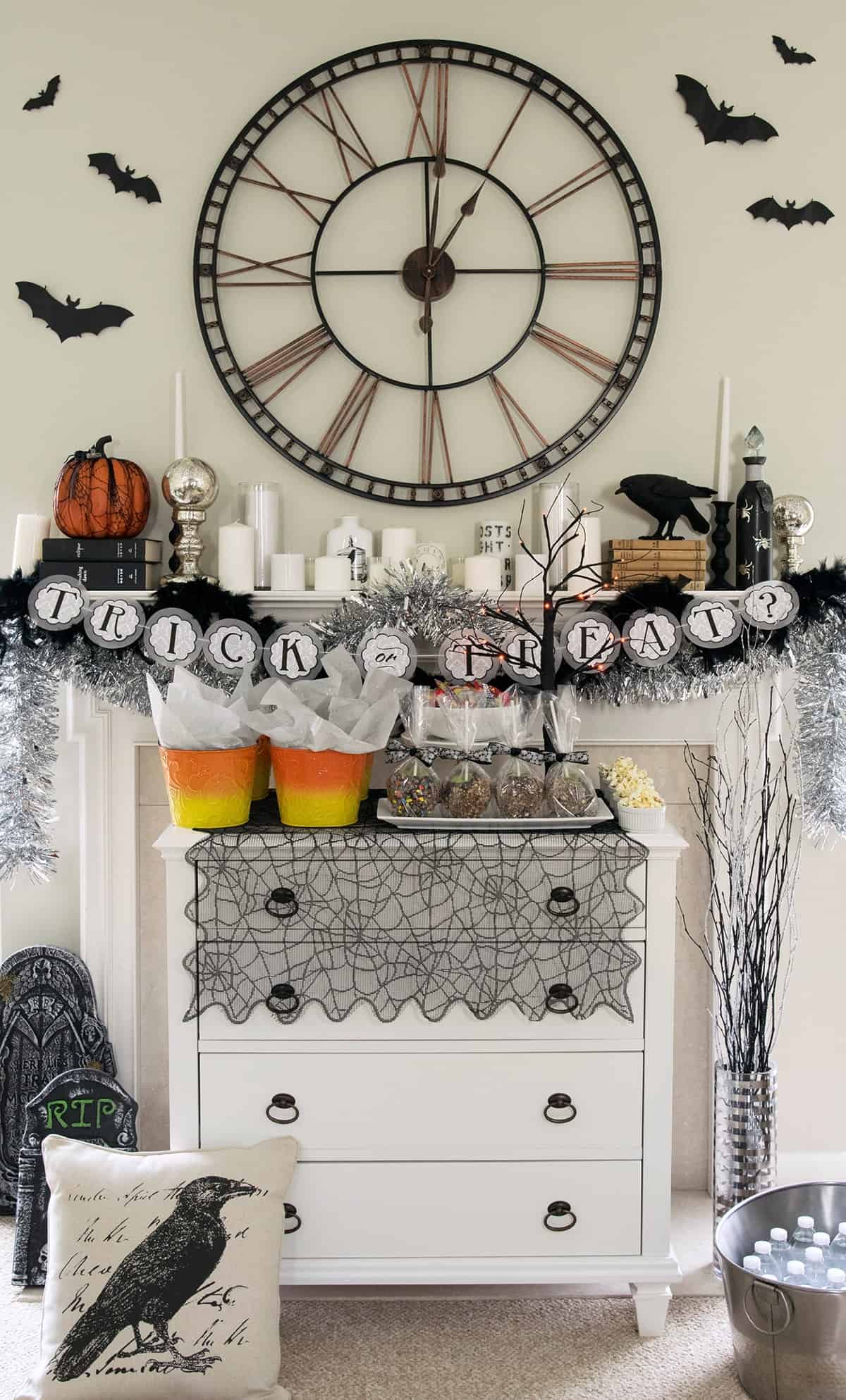 Halloween Party Decorations on a mantle with bats, an old time clock, candles, books, and an assortment of goodies on a buffet table.