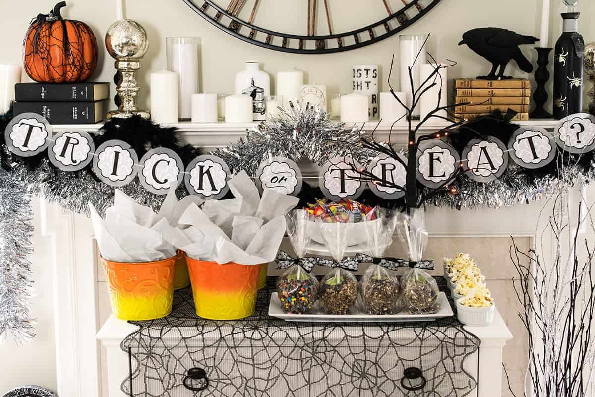 Halloween buffet table with layered decorations and a trick or treat banner. Table has candy corn buckets and apples on it.