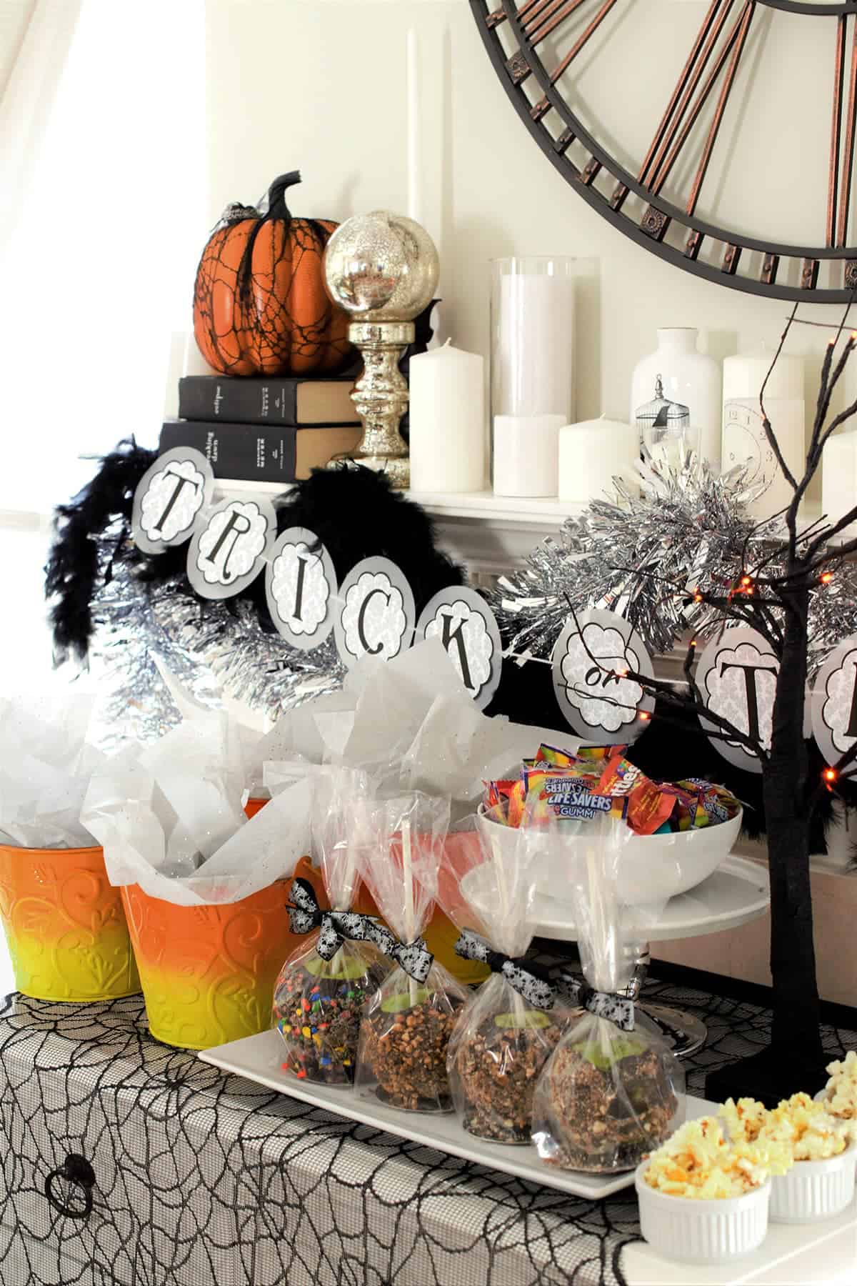 Halloween decorations on a white mantle and treat table for entertaining guests in front and full of treats like candy, popcorn, and dipped apples.