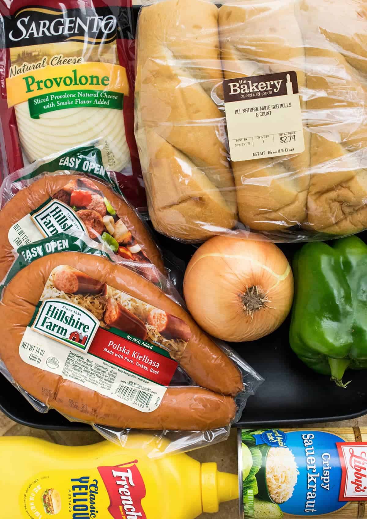 Ingredients for polish sausage sandwiches including kielbasa, hoagie rolls, peppers, onions, slices provolone, sauerkraut, and mustard.