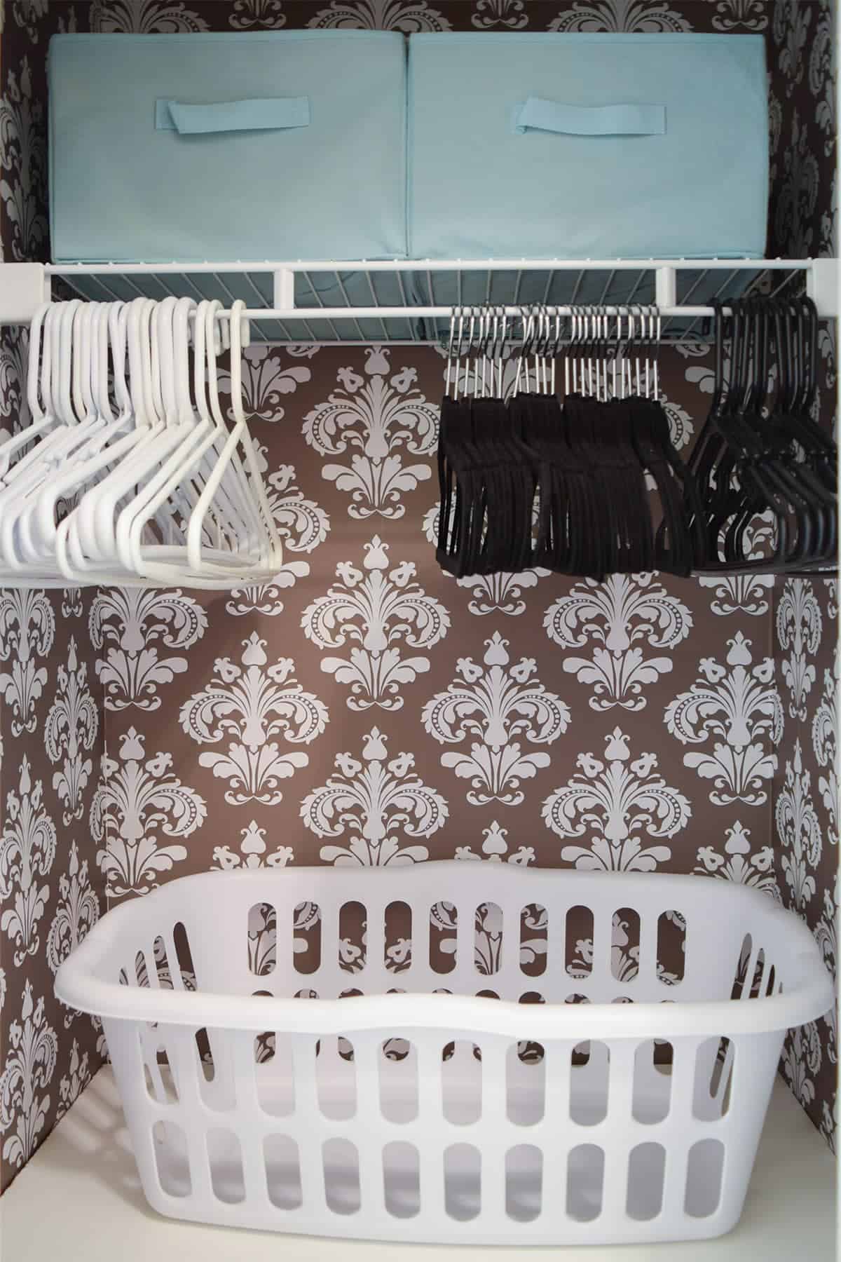 Small laundry closet with brown and white damask wall covering, white shelves with laundry baskets, black and white hangers, and teal bins.