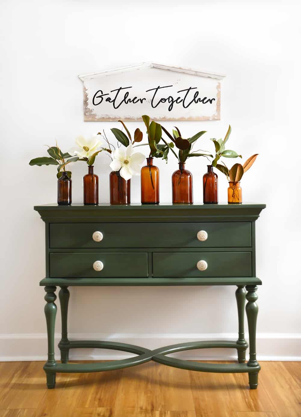 Lush painted deep green console table furniture with round hardware. Collection of brown glass bottle vases filled with magnolia blossoms on top.