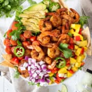 Overhead of Mango Shrimp Salad in a bowl with sliced avocado, jalapeno, grape tomatoes, mango, peppers, lime, cilantro and tortilla chips with bottle of dressing on the side.