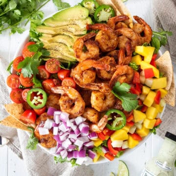Overhead of Mango Shrimp Salad in a bowl with sliced avocado, jalapeno, grape tomatoes, mango, peppers, lime, cilantro and tortilla chips with bottle of dressing on the side.