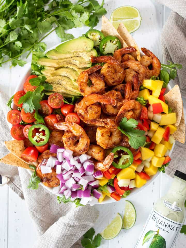 Overhead of Mango Shrimp Salad in a bowl with sliced avocado, jalapeno, grape tomatoes, mango, peppers, lime, cilantro and tortilla chips with bottle of dressing on the side.
