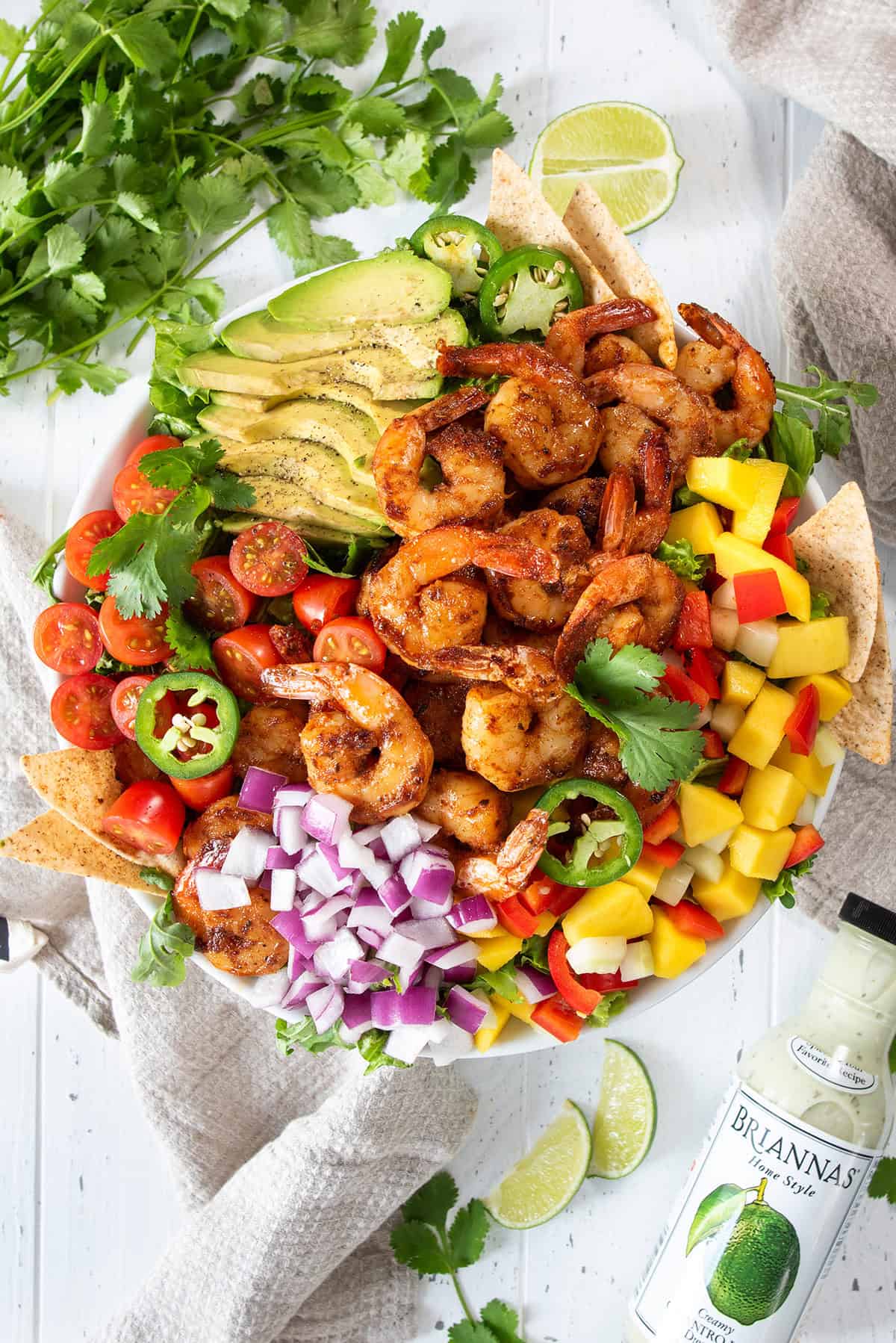 Overhead of Mango Shrimp Salad in a bowl with sliced avocado, jalapeno, grape tomatoes, mango, peppers, lime, cilantro and tortilla chips with bottle of dressing on the side.