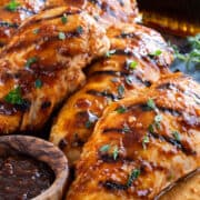 Several grilled chicken breasts with sauce on a wood board with a bottle of maple syrup.