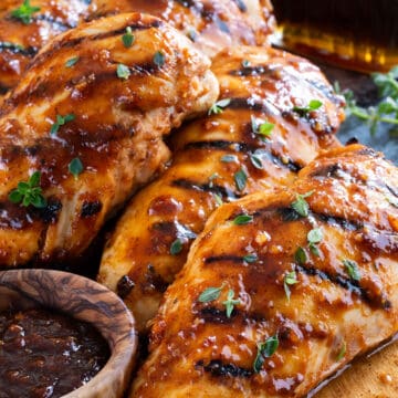 Several grilled chicken breasts with sauce on a wood board with a bottle of maple syrup.